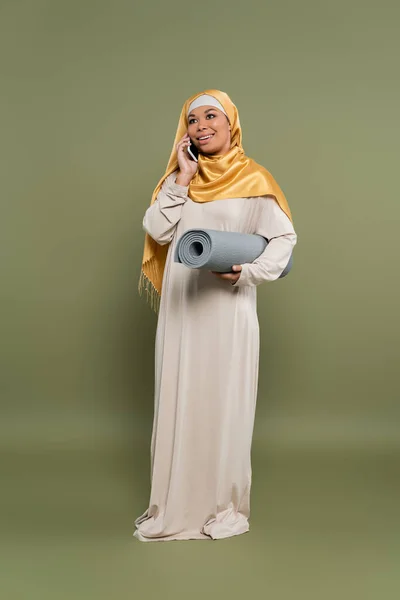 Full length of multiracial woman in hijab holding fitness mat and talking on smartphone on green background — Stock Photo