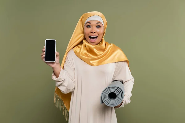 Excited multiracial woman in hijab holding smartphone and fitness mat on green background — Stock Photo