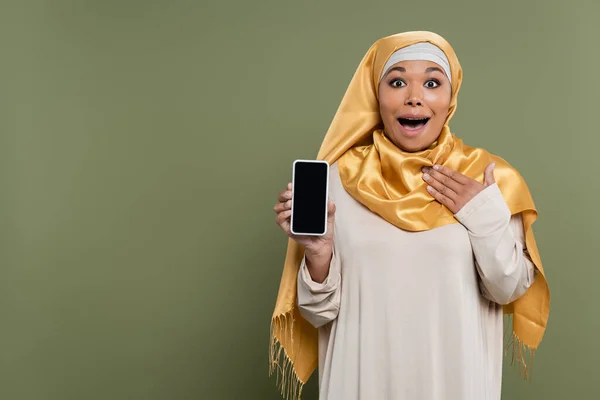 Excited multiracial woman in hijab holding cellphone with blank screen on green background — Stock Photo