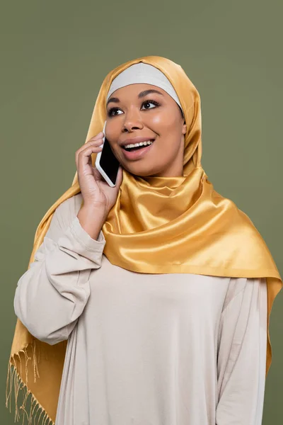 Sonriente mujer multirracial en hijab hablando en smartphone aislado en verde - foto de stock