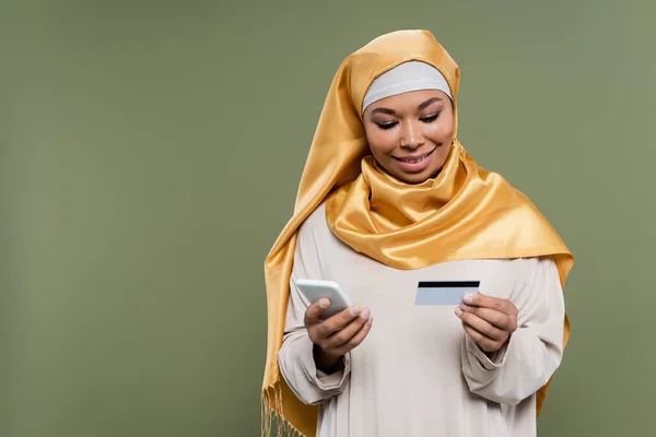 Smiling multiracial woman in hijab using smartphone and credit card isolated on green — Stock Photo