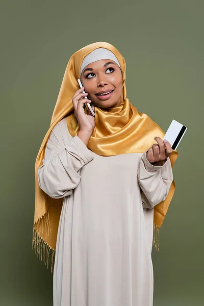 Позитивная многорасовая женщина в хиджабе разговаривает на смартфоне и держит кредитную карту изолированы на зеленый — стоковое фото
