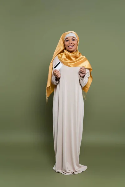 Полная длина улыбающейся многорасовой женщины в хиджабе держа кредитную карту на зеленом фоне — стоковое фото