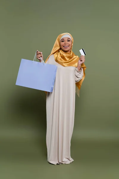 Cheerful multiracial woman in hijab holding credit card and shopping bag on green background — Stock Photo