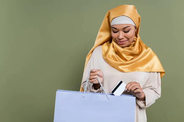 Multiracial woman in hijab putting credit card in shopping bag isolated on green — Stock Photo