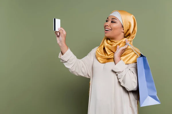 Cheerful multiracial woman in golden hijab holding credit card and shopping bag on green background — Stock Photo