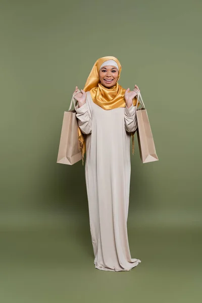 Mujer multirracial positiva en hijab sosteniendo bolsas de compras sobre fondo verde - foto de stock