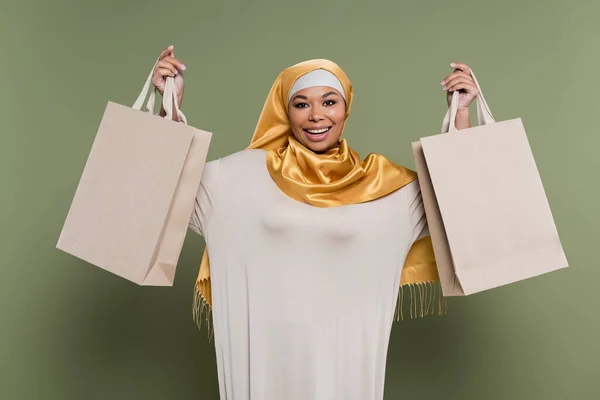 Femme multiraciale joyeuse en hijab jaune tenant des sacs à provisions sur fond vert — Photo de stock