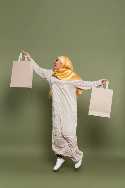 Vista lateral de la mujer musulmana multirracial emocionada sosteniendo bolsas de compras y saltando sobre fondo verde - foto de stock