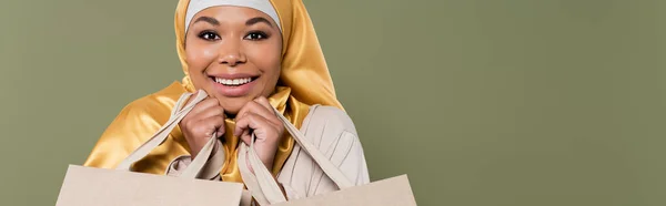 Cheerful multiracial woman in golden hijab holding shopping bags isolated on green, banner — Stock Photo