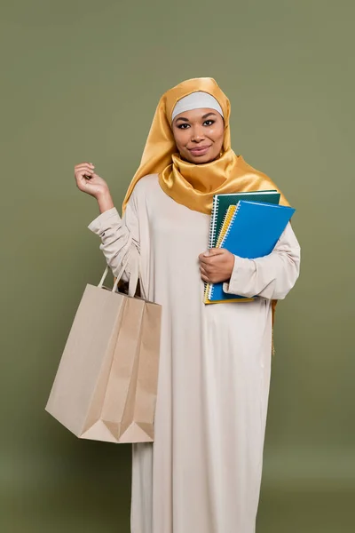 Young multiracial woman in hijab holding shopping bags and notebooks on green background — Stock Photo