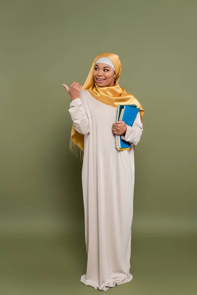 Lächelnder multikultureller Student im Hijab hält Notizbücher in der Hand und zeigt Daumen nach oben auf grünem Hintergrund — Stockfoto