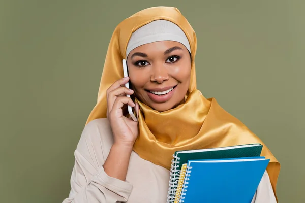 Retrato de estudiante multirracial en hijab hablando en smartphone y sosteniendo cuadernos aislados en verde - foto de stock
