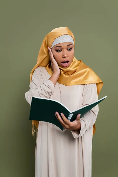 Shocked multiracial student in hijab holding notebook on green background — Stock Photo