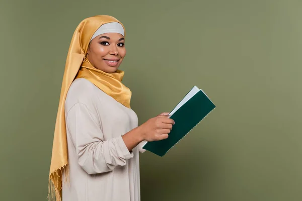 Positive multikulturelle Studentin im Hijab hält Notizbuch in der Hand und blickt in die Kamera auf grünem Hintergrund — Stockfoto