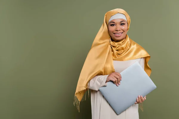 Positiva donna multirazziale in hijab in possesso di laptop e guardando la fotocamera su sfondo verde — Foto stock