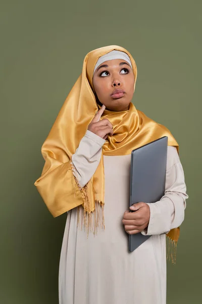 Pensive multiracial woman in hijab holding laptop on green background — Stock Photo