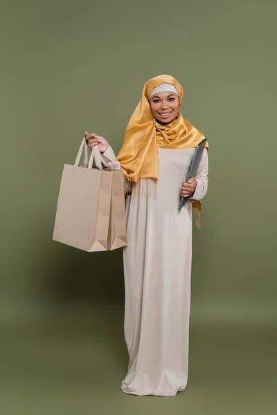 Longitud completa de la mujer multicultural en hijab celebración de la computadora portátil y bolsas de compras sobre fondo verde - foto de stock