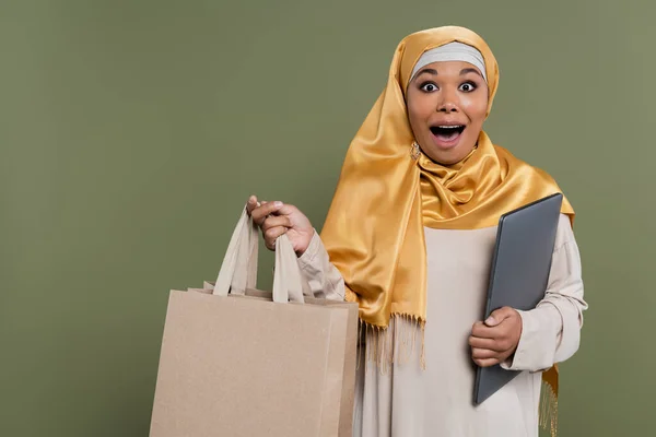 Excited multicultural woman in hijab holding laptop and shopping bags isolated on green — Stock Photo