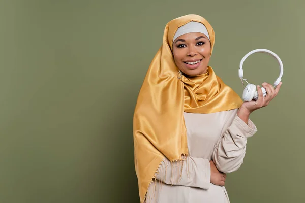 Mujer multirracial sonriente en hijab sosteniendo auriculares sobre fondo verde - foto de stock