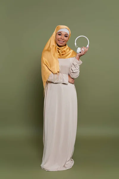 Повна довжина позитивної мультикультурної жінки в хіджабі, що тримає бездротові навушники на зеленому фоні — стокове фото