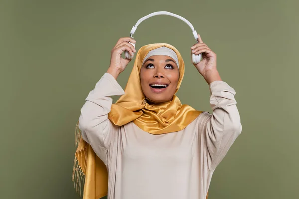 Позитивная мультикультурная женщина в хиджабе держит беспроводные наушники изолированы на зеленый — стоковое фото