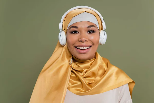 Joven mujer multirracial en hijab y auriculares mirando a la cámara aislada en verde - foto de stock