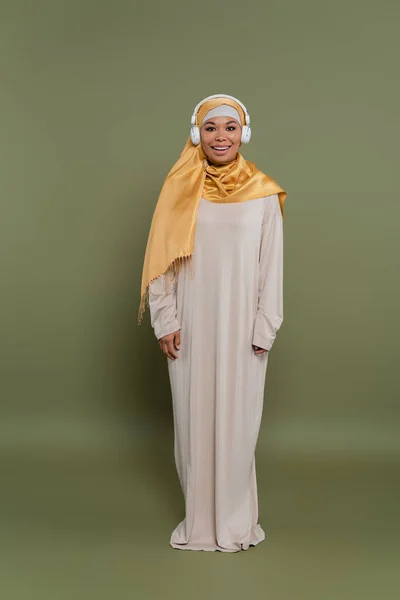Volle Länge der positiven multirassischen Frau im Hijab und Kopfhörer auf grünem Hintergrund — Stockfoto