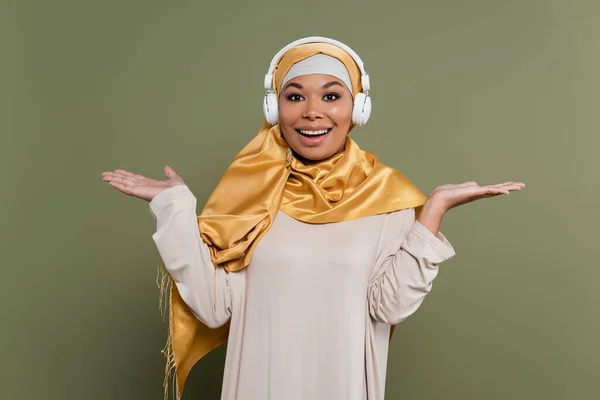 Positive multiracial woman in hijab and headphones pointing with hands on green background — Stock Photo