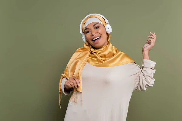 Веселая многорасовая женщина в хиджабе и наушниках поет на зеленом фоне — стоковое фото