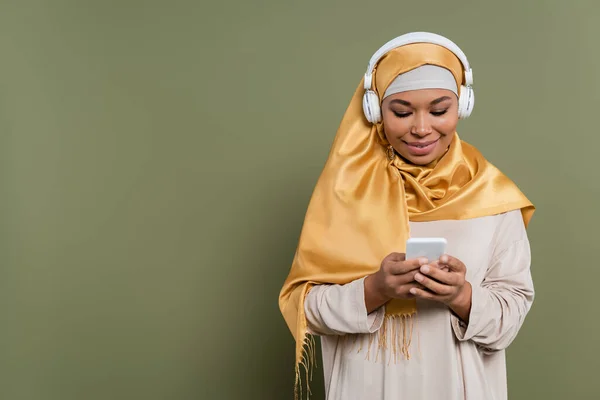Smiling multiracial woman in hijab and headphones using smartphone on green background — Stock Photo
