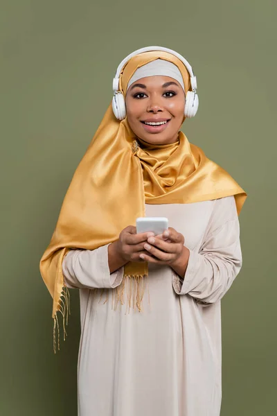 Mujer multirracial positiva en hijab y auriculares que sostienen el teléfono inteligente sobre fondo verde - foto de stock