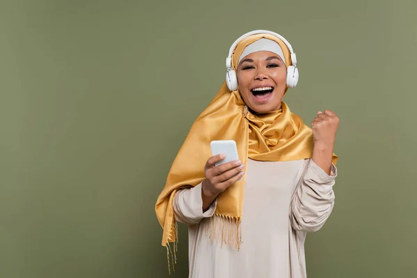Excited multiracial woman in hijab and headphones holding smartphone on green background — Stock Photo