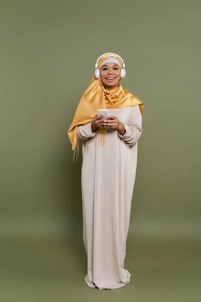 Fröhliche Frau in Hijab und Kopfhörer mit Handy auf grünem Hintergrund — Stockfoto