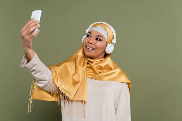 Lächelnde multiethnische Frau im Hijab und Kopfhörer, die ein Selfie mit dem Smartphone auf grünem Hintergrund macht — Stockfoto