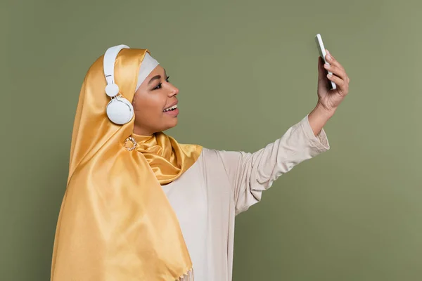 Позитивная многорасовая женщина в хиджабе и наушниках делает селфи на мобильном телефоне, изолированном на зеленом — стоковое фото