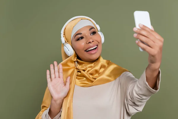 Femme multiraciale joyeuse en hijab et écouteurs ayant appel vidéo sur smartphone isolé sur vert — Photo de stock