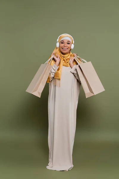 Mujer alegre multirracial en auriculares y hijab sosteniendo bolsas de compras sobre fondo verde - foto de stock