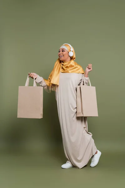 Улыбающаяся многорасовая женщина в хиджабе и наушниках, держащая сумки с покупками и идущая на зеленом фоне — стоковое фото