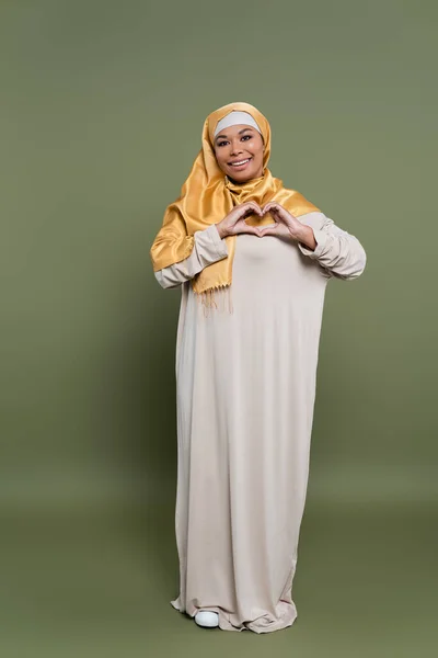 Mulher multirracial despreocupada no hijab mostrando sinal de amor com as mãos no fundo verde — Fotografia de Stock