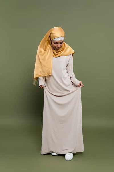 Стильная многорасовая женщина в хиджабе, стоящая в платье на зеленом фоне — стоковое фото