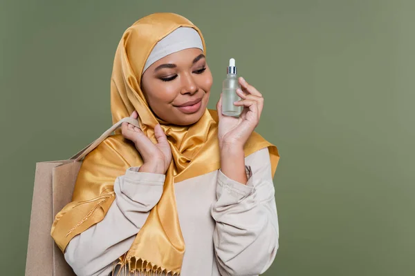 Smiling multiracial woman in hijab holding shopping bag and serum isolated on green — Stock Photo