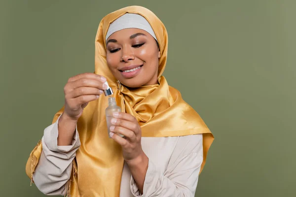 Positive multiracial woman in hijab holding cosmetic serum and dropper isolated on green — Stock Photo