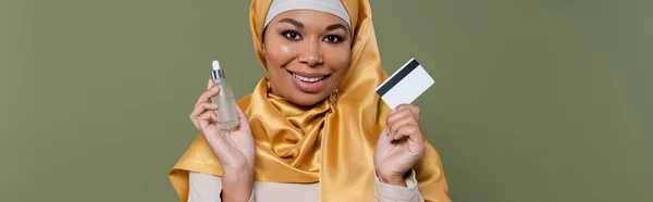 Cheerful multiracial woman in hijab holding credit card and serum isolated on green, banner — Stock Photo