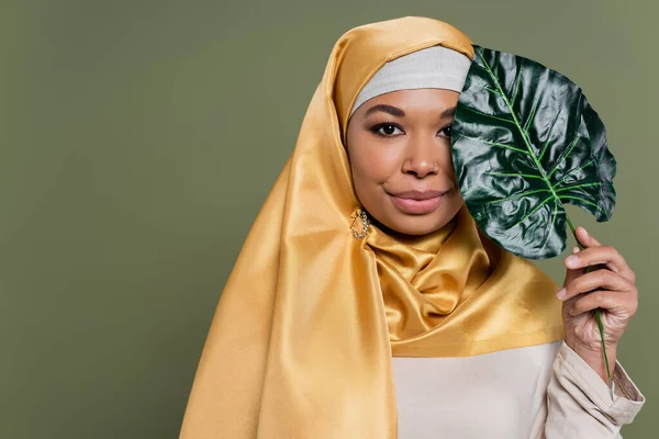Young multiracial woman in hijab holding tropical leaf isolated on green — Stock Photo