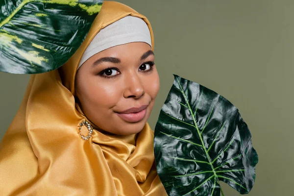 Retrato de mulher multirracial no hijab olhando para a câmera perto de folhas isoladas no verde — Fotografia de Stock