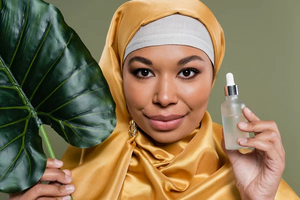 Portrait de femme musulmane multiraciale en hijab de soie jaune tenant sérum cosmétique et feuille tropicale isolée sur vert — Photo de stock