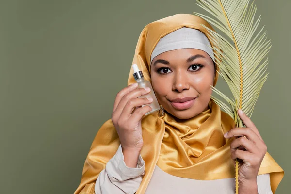 Mujer musulmana multirracial en hiyab satinado amarillo sosteniendo hoja tropical y botella de suero cosmético aislado en verde - foto de stock