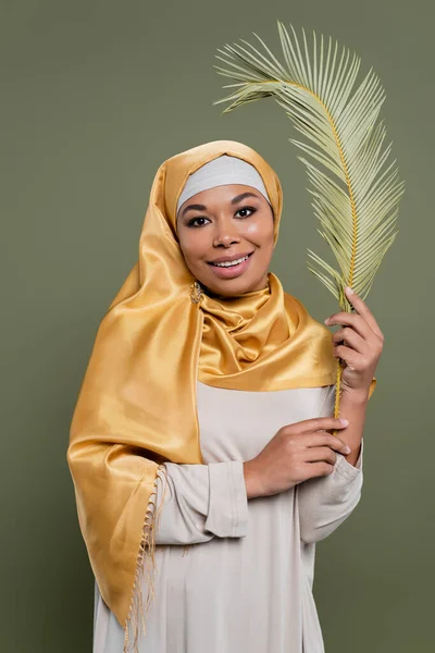 Alegre mujer multirracial en amarillo hijab de seda sosteniendo hoja de planta exótica y sonriendo a la cámara aislada en verde - foto de stock