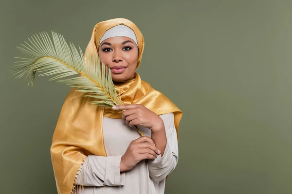 Mujer multirracial feliz con maquillaje usando hiyab satinado amarillo y sosteniendo hoja exótica aislada en verde - foto de stock